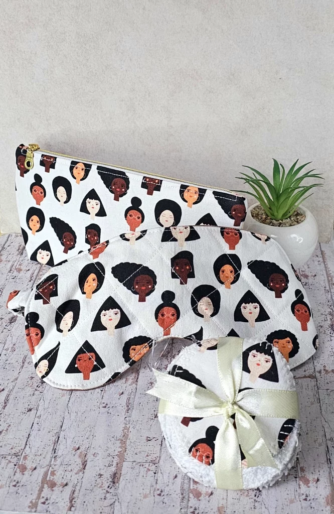 Self care gift set of a toiletry bag and matching sleep mask made in fabric print featuring illustrated heads of women of different cultures on a white background.